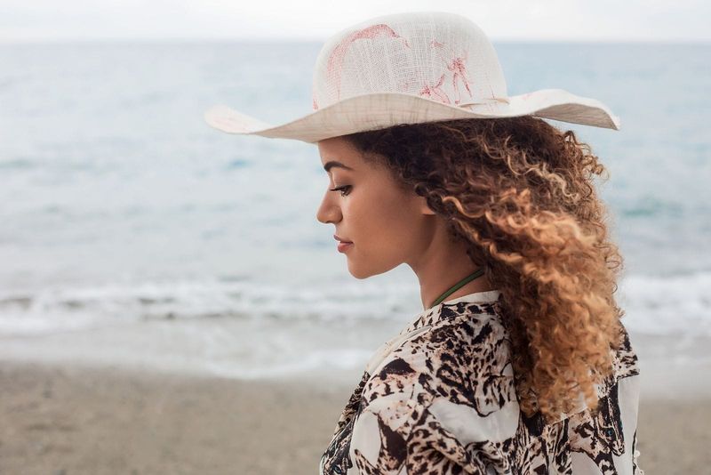 donna splendida pensosa in piedi vicino al mare che indossa un cappello