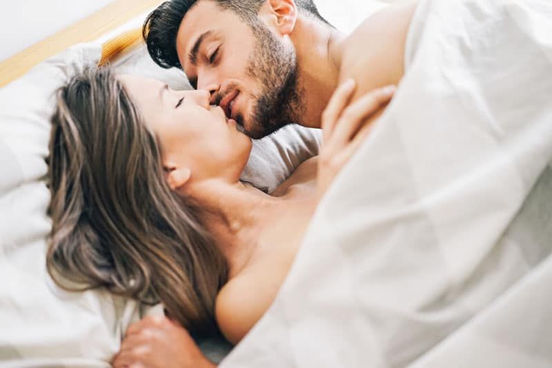 người đàn ông mỉm cười hôn người phụ nữ dưới ga trải giường