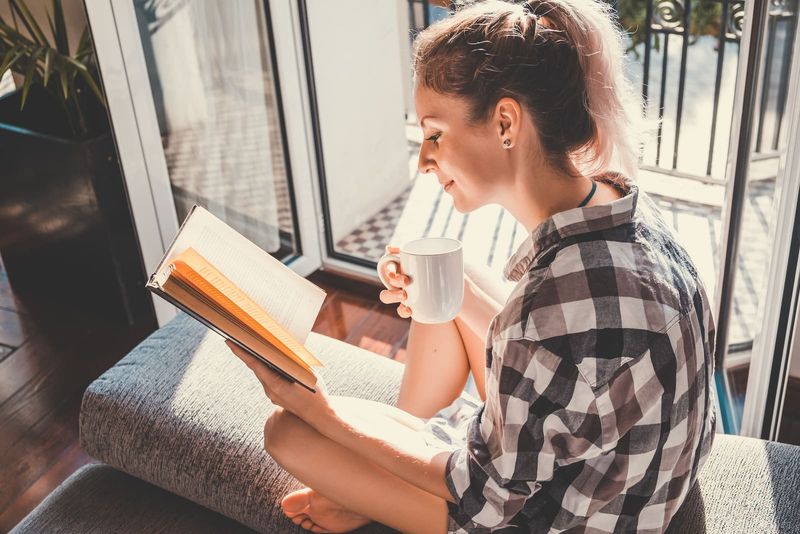 Tânără femeie drăguță așezată la fereastra deschisă, bea cafea și citește o carte