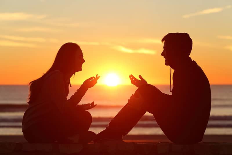 صورة ظلية لزوجين يتحدثان في غروب الشمس أثناء الجلوس بجانب الماء