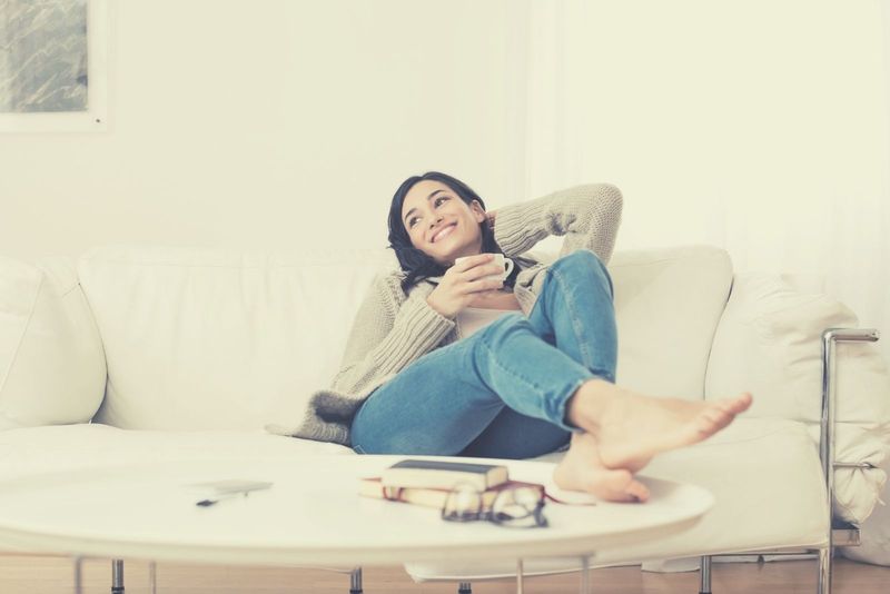 người phụ nữ mỉm cười suy nghĩ và nằm dài trên ghế sofa trong phòng khách và cầm một chiếc cốc