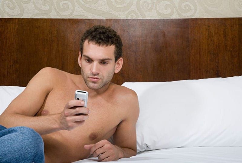 home en topless enviant missatges de text al llit amb llençols blancs