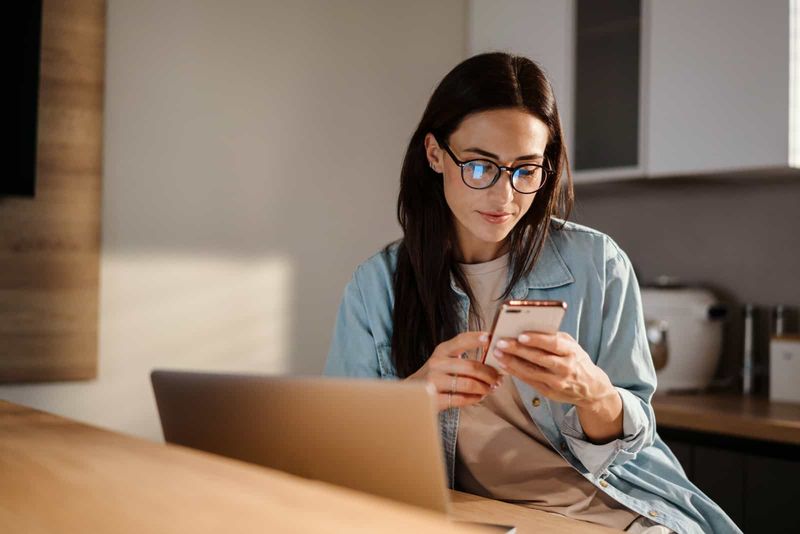 eine schöne frau mit langen schwarzen haaren mit brille, die an einem tisch sitzt und auf ein handy drückt