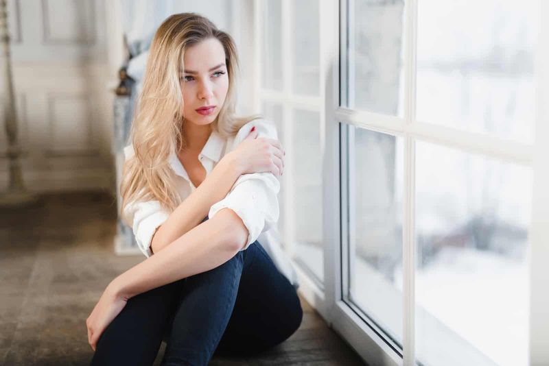 przy oknie siedzi smutna kobieta z długimi blond włosami