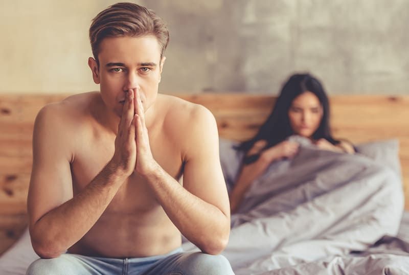 un bărbat serios stând pe marginea patului în timp ce prietena lui stă întinsă în pat