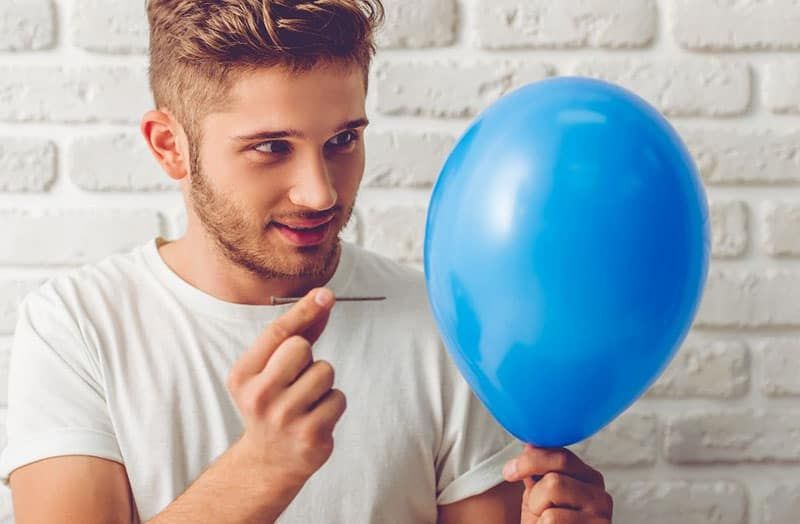 pria yang akan meledakkan balon biru dengan jarum di dekat dinding bata putih