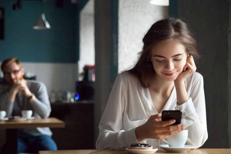 امرأة تحدق في هاتفها الذكي وهي تبتسم داخل المقهى مع رجل في الخلف يحدق بها