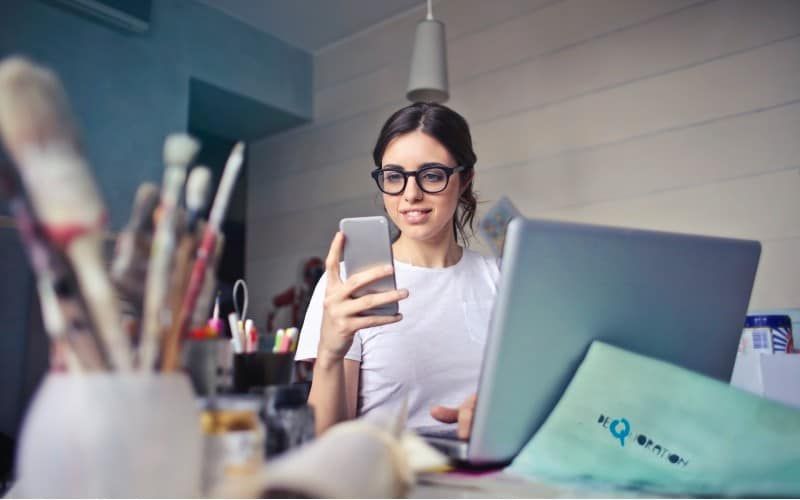 kobieta w białej koszuli używająca smartfona siedząca przy biurku z laptopem na nim
