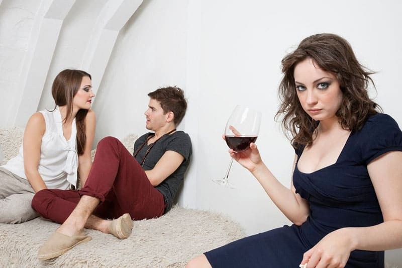 Eifersüchtige Frau, die Wein nippt, entfernt von einem süßen Pärchen, das sich unterhält