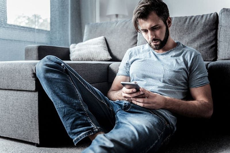 cheerles trauriger mann, der auf sein handy schaut und auf dem boden neben dem sofa sitzt