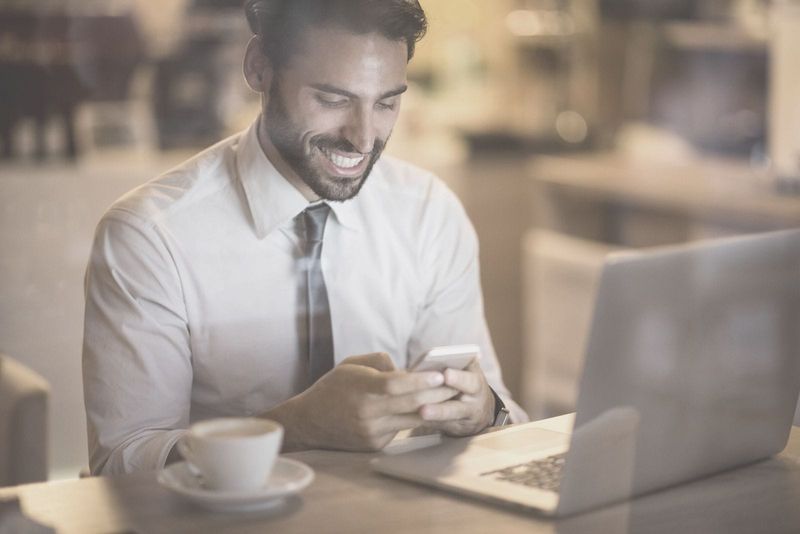 bärtiger mann lächelt, liest nachrichten von seinem telefon in einem café mit einem laptop und kaffee auf dem tisch