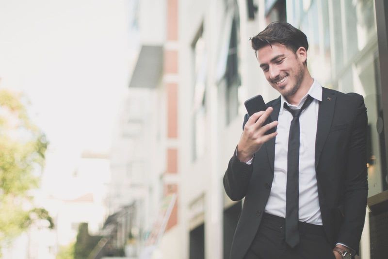 Lächelnder Geschäftsmann SMS mit seinem Telefon an die Wand gelehnt im Freien