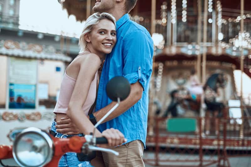 người đàn ông mặc áo sơ mi xanh đang ôm người phụ nữ trẻ tóc vàng đang mỉm cười bên ngoài