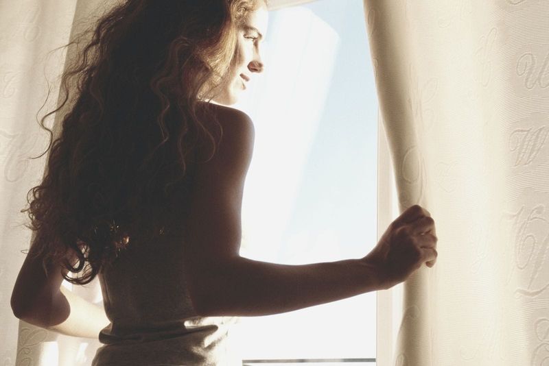 mlada žena koja otvara prozor i smiješi se ujutro sa zrakama sunca koje je udaraju