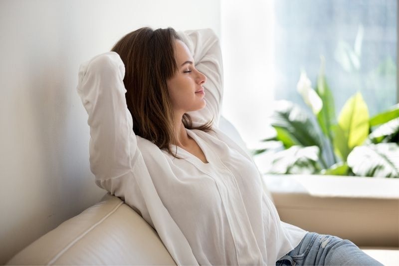 wanita yang tenang duduk di sofa di ruang tamu menutup matanya dengan tangan di belakang kepalanya