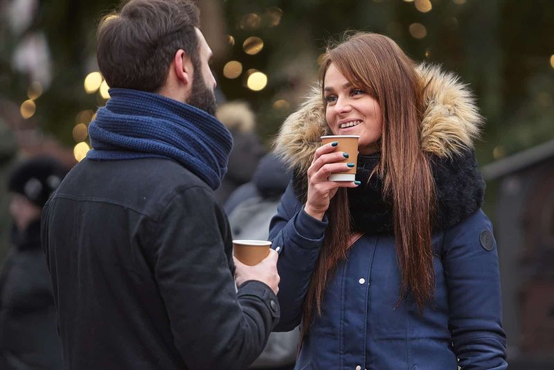 un bărbat și o femeie zâmbitori stând în aer liber ținând cești de cafea în mână și vorbind