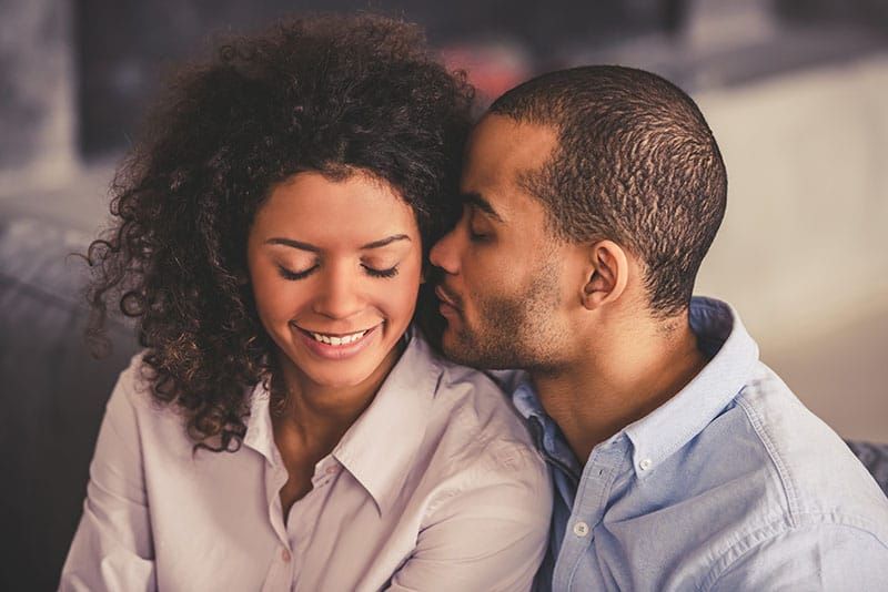美しい若いアフロアメリカ人のカップルは、自宅のソファに一緒に座っている間、抱き合って笑っています