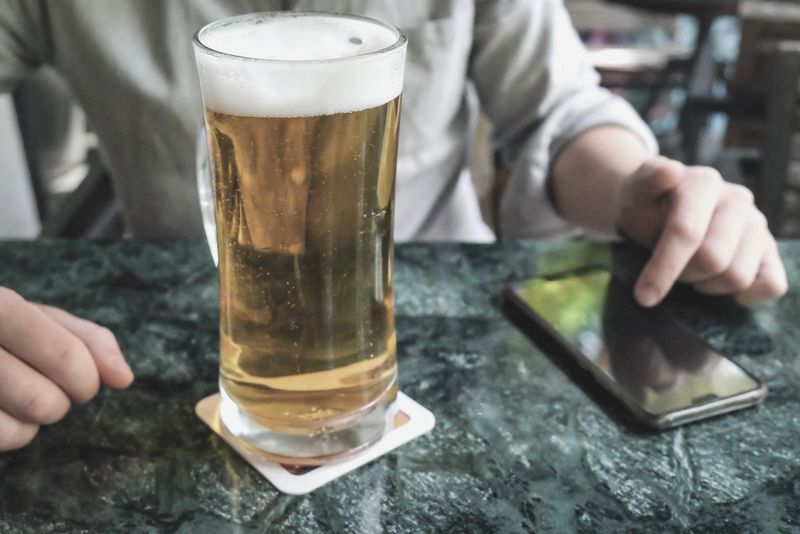 um copo de cerveja colocado na mesa com uma imagem recortada de uma mulher pressionando as teclas do smartphone em cima da mesa