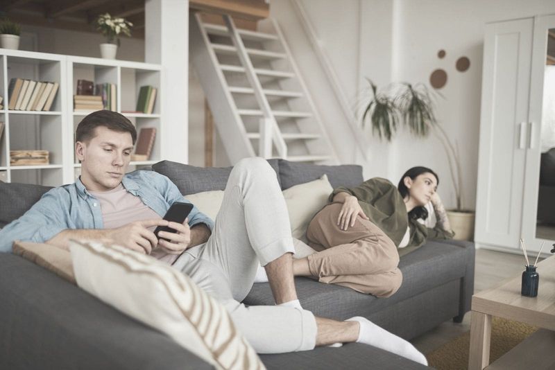 casal entediado em casa assistindo tv no sofá com foco no homem ocupado com seu gadget