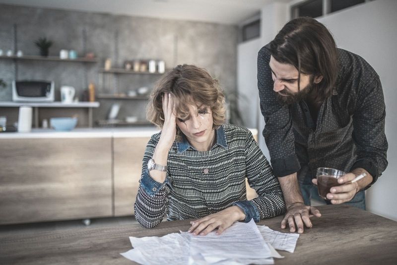 casal com problemas financeiros com a mulher chorando olhando as contas e o homem segurando uma cerveja e um charuto dentro da cozinha