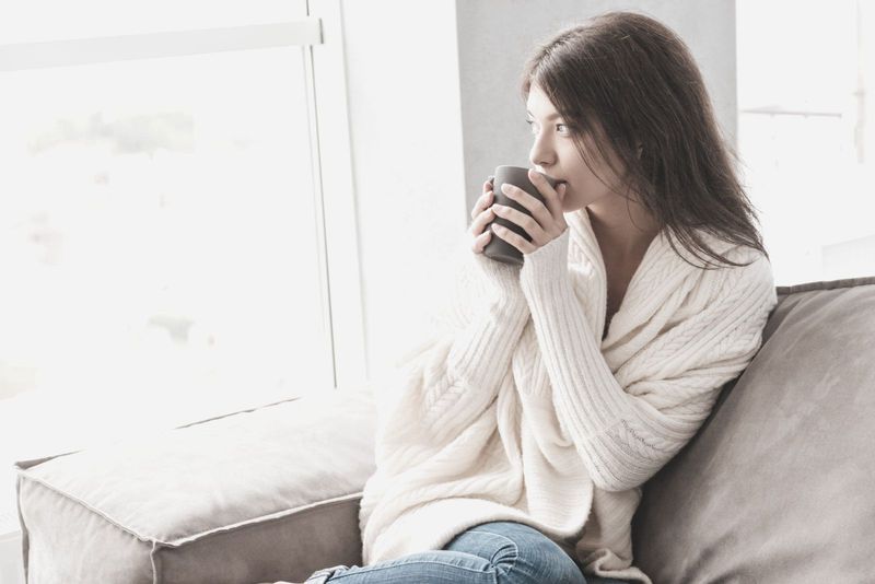 mulher jovem e pensativa tomando seu café no início da manhã descansando no sofá perto das janelas