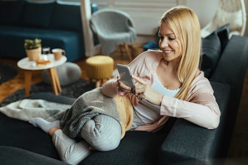 seorang wanita dengan rambut pirang panjang duduk di sofa dan memegang telepon di tangannya