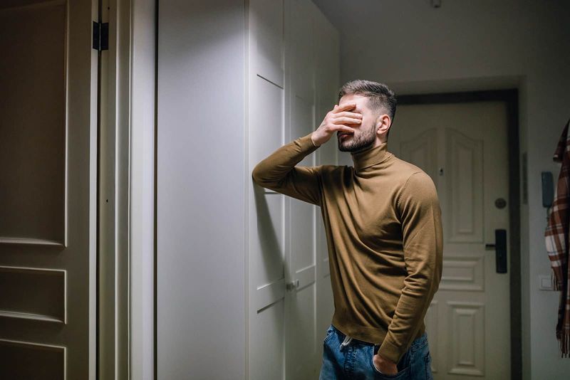 een trieste man die zijn gezicht bedekt terwijl hij tegen de muur leunt in een appartement