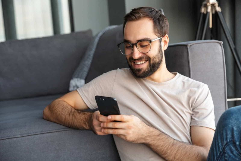 una mujer sonriente sentada en el suelo apoyada en el sofá y presionando un botón en el teléfono