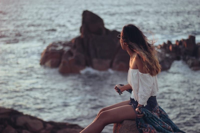 mulher de top branco sentado na pedra olhando para o mar