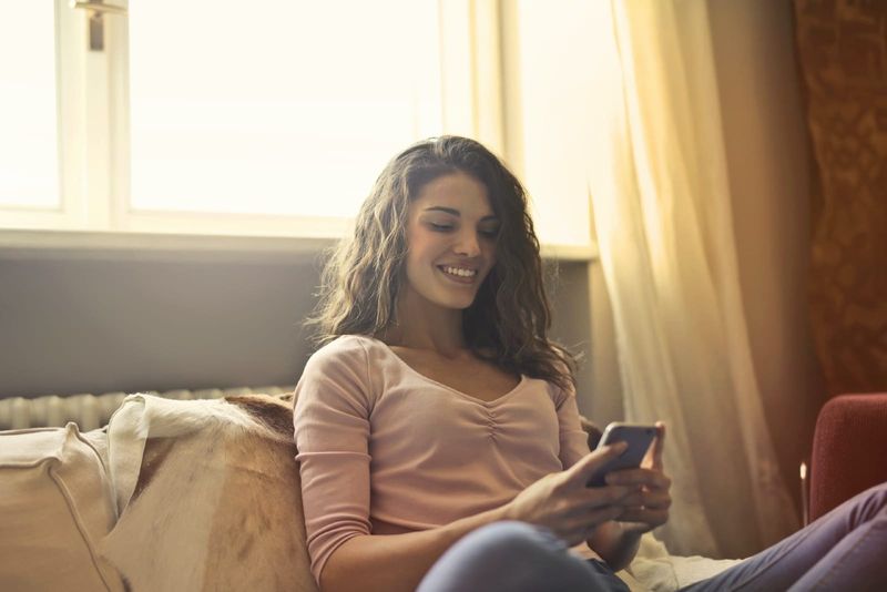 glückliche frau, die auf das telefon schaut, während sie auf dem bett sitzt