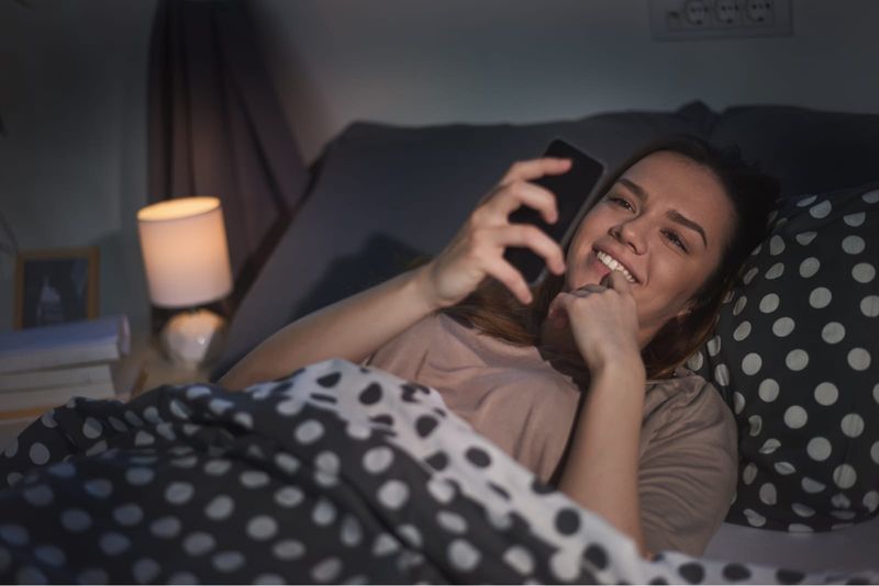 seorang gadis bahagia di malam hari berbaring di tempat tidur dan menggunakan ponsel