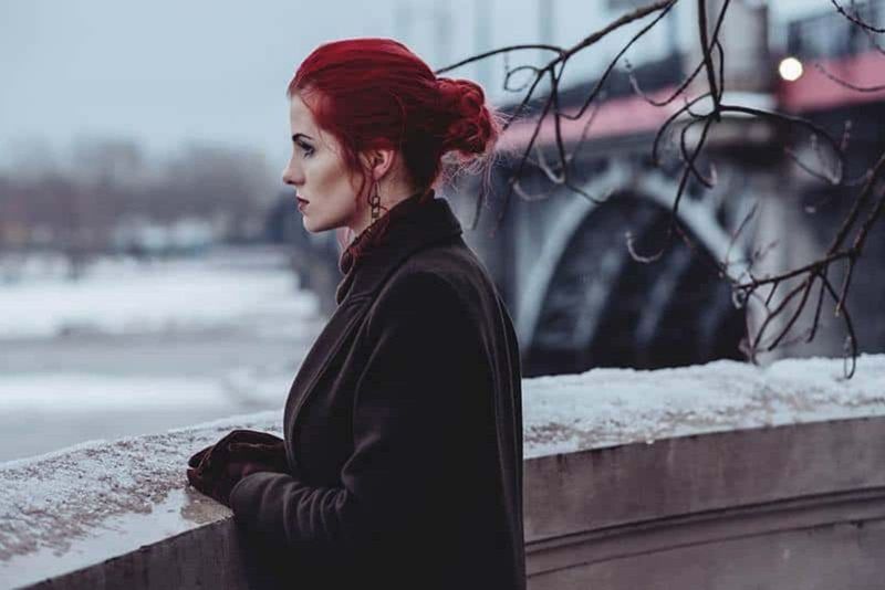 mulher pensativa com cabelo vermelho e jaqueta marrom do lado de fora