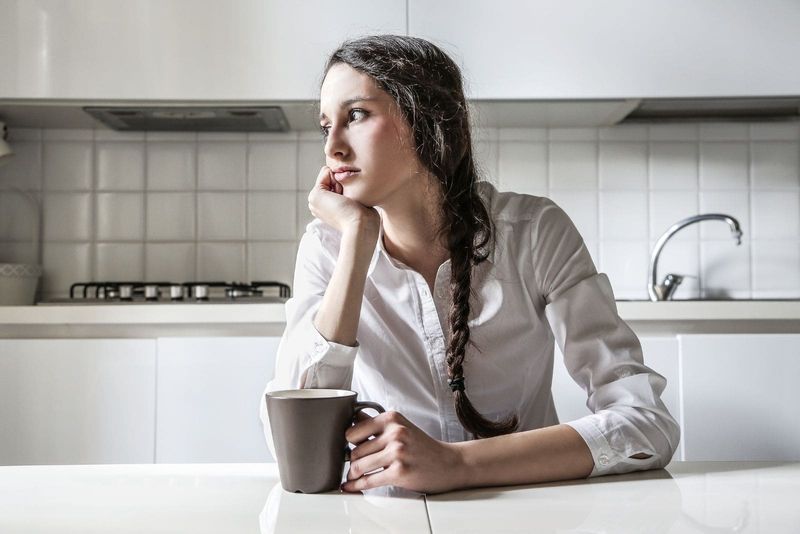 femeie gânditoare care bea cafea în bucătărie privind în altă parte