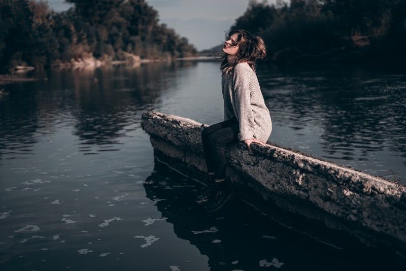 žena u sivom džemperu sjedi na betonu u blizini vode