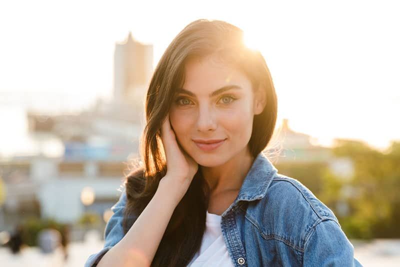 Porträt einer schönen lächelnden jungen Brunettefrau, die draußen mit Stadtbildhintergrund steht und aufwirft