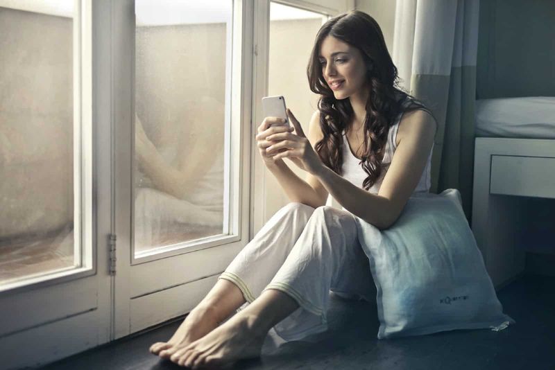 Wanita bahagia menggunakan smartphone sambil duduk di dekat jendela
