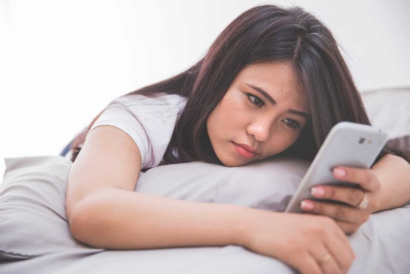 wanita sedih berbaring di tempat tidur sambil menggunakan smartphone